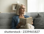 Small photo of Dreamy pensive senior reader woman sitting on cozy couch at home, reading book, bestseller novel, looking away, thinking over story, feeling lazy, peace, enjoying literature hobby