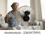 Small photo of Close up electric ventilator with propeller cooling air blowing to senior older lady suffering from overheating, hypoxia, sitting on sofa with closed eyes, flying hair in blurred background