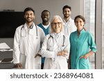 Small photo of Positive confident senior clinic chief doctor and younger diverse team of practitioners, surgeons, nurses standing together in hospital office, looking at camera with toothy smiles