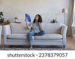 Small photo of Exhausted frustrated Hispanic woman waving paper handheld fan, cooling hot air, suffering from heat attack, headache, hypoxia, stuffy air, sitting on home couch in living room