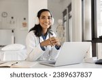 Small photo of Cheerful beautiful young doctor woman in head phones posing at hospital workplace with laptop computer, looking at camera with toothy smile, offering online medical consultation. Professional portrait