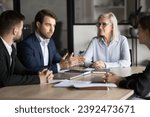 Small photo of Confident younger project manager man in formal suit speaking to colleagues and elder leader at meeting table, explaining work plan, teamwork strategy, giving report, ideas for brainstorming