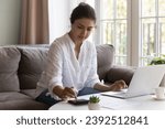 Small photo of Serious Indian woman sit on sofa use calculator calculate personal expenses, pay utility bills via e-bank app on laptop, manage budget, control outcomes and incomes, do accounting work at home concept