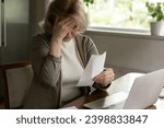 Small photo of Financial problem. Stressed confused older woman sit by work desk at home office touch forehead look at overdue bill tax debt notification. Worried mature businesswoman has to bear extra costs taxes