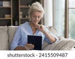 Small photo of Serious mature female using digital tablet, sit on sofa with device, read on-line news, learning new application looks concerned, due to lack of understanding. Older gen and modern wireless tech usage