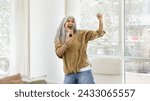 Small photo of Cheerful excited elderly Hispanic woman singing at supposed microphone at home, pretending celebrity, playing singer, dancing in apartment interior, enjoying entertainment, funny activity. Banner shot