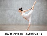 Profile portrait of beautiful young woman wearing white sportswear working out against grey wall, doing yoga or pilates exercise. Standing in variation of Natarajasana, Lord of the Dance. Full length