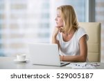 Young attractive woman at a modern office desk, working with laptop, looking at the window, thinking about a post, full-time blogger, seeking for inspiration, help to be productive, updating computer