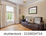 Small photo of Burrough Green, Suffolk - July 12 2018: Small living room snug with leather sofa and window dressed with floor to ceiling curtains
