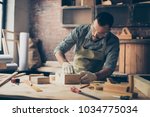 Small photo of Concentrated talented serious confident bearded handicraftsman wearing casual checkered shirt apron gloves and safety glasses is polishing a wooden box using sandpaper