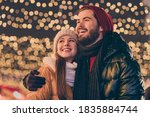 Photo of affectionate boyfriend hug girlfriend look enjoy x-mas tradition, illumination evening outside wear season coat scarf headwear
