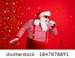 Photo of grandpa grey beard hold mic open mouth scream sing song karaoke wear santa claus x-mas costume suspenders sunglass striped shirt cap isolated red color background