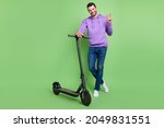 Full body photo of young cheerful man happy positive smile ride scooter show peace v-sign isolated over green color background