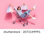 Photo of cute excited young woman wear white sweater sitting shopping cart holding bargains smiling isolated pink color background