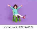 Small photo of Full length photo of excited funky small kid wear turquoise shirt sitting chair rising hands arms empty space isolated violet color background