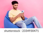 Small photo of Photo of young concentrated man with wavy hair using his apple iphone last model sit beanbag rest time isolated on pink color background