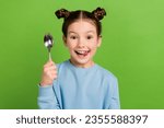 Small photo of Portrait of little girl with brown buns hairstyle wear blue pullover lick teeth hold spoon wait dinner isolated on green color background