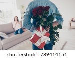 Unexpected moment in routine everyday life! Cropped photo of man's hands hiding holding chic bouquet of red roses and gift with white ribbon behind back, happy woman is on blurred background