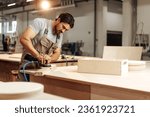 Young carpenter sanding wood piece in workshop in furniture factory