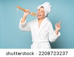 Portrait of woman singer taking high note singing in massage brush on blue studio background dressed in white bathrobe and head towel. Beauty and spa, morning bath