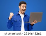 We got a winner! Excited young man in blue shirt looking at laptop screen with WOW expression, isolated on blue background