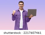 He's a winner! Happy young man in purple shirt looking at laptop screen with victory expression, isolated on purple background