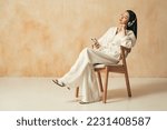 Side view of pretty asian woman sitting on chair on brown copy space background in home clothes, listening music in wireless headphones with closed eyes, using audio player app for phone