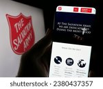Small photo of Stuttgart, Germany - 10-09-2023: Person holding smartphone with website of Protestant charity organization The Salvation Army (TSA) with logo. Focus on center of phone display. Unmodified photo.