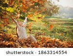 Autumn leaves falling on happy young woman in forest. The colors and mood of autumn. Beautiful young woman throwing leaves in a park. Fall concept. Happy smiling girl with natural red hair