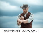 Small photo of Cowboy farmer man in country side wearing western cowboy hat. American Male model in countryside on farm. Cowboy with lasso rope on sky background.