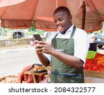 Small photo of A happy African male trader with a smart phone, standing beside his stall of tomatoes and pepper in a market place