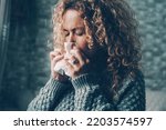 Small photo of Side view of woman blowing nose for flu influenza symptoms in winter at home. Temperature reduction indoor to save energy gas costs. Female people with virus contagion using paper tissues. Disease