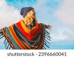 Small photo of Blonde woman in hat and colorful poncho relaxing with open arms in blue winter sky. People's freedom style. People freedom style. Traveler outstretching arms outdoor leisure activity