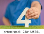 Small photo of Number 4 in hand. Hand holding white number four over green grass and purple background, birthday concept, competition place, four percent per annum. Canada National Day, USA Independence Day, 4 July.