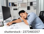 Young adult Asian businessman sleeping on his desk with a computer pc in the office setting background. - Workaholic, boring, exhausted, sleepy business man concept