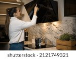 An adult woman using the control panel of the modern cooker hood. Kitchen room with cozy interiors and contemporary built-in appliances.
