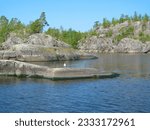Rocky coast with large stones, trees and reflection in quiet water surface of northern lake. Northern nature and blue sky in summer. Ladoga lake, Karelia, Russia.