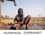 A young African girl casually crouches in front of a water pump and washes roughly, which is symbolic of the lack of tap water and washing facilities in the developing world