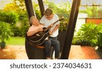 Small photo of Happy mature middle aged man guitarist sitting on chair play acoustic guitar enjoy music outdoor home. Beautiful smiling Indian female singing practice tuning on musical instrument spend time together