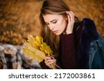 A sad beautiful girl with red long hair in warm sweater and jeans coat holds a bouquet of autumn yellow maple leaves. Melancholic mood, autumn vybe, selective focus. High quality photo.