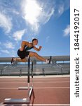 Small photo of Side view of a male athlete jumping over a hurdle in the race on running track