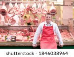 A young smiling butcher in a red apron standing and leaning his back against the butcher shop window.