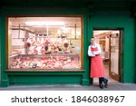 A wide shot of a young smiling butcher in red apron leaning against butcher shop doorway next to the display window.