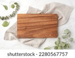 Small photo of Wood cutting board on linen napkin with leaves on marble background, top view