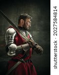 Small photo of Oath to the king. Portrait of one brutal bearded man, medeival warrior or knight with dirty wounded face holding sword and shield isolated over dark background.