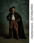 Small photo of Portrait of genteel brutal man, medeival pirate in hat and cloak with sword isolated over dark background. Combination of medeival and modern styles. Concept of history. Copyspace for ad.