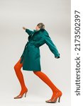 Small photo of Side view portrait of stylish girl in green coat and bright orange tights dancing isolated over grey background. Expressive fashion. Concept of retro fashion, art photography, style, queer, beauty