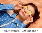 Strange look. Weird people concept. Young girl lying on table with eggs on her eyes over peach color background. Vintage, retro style. Food pop art photography, fashion, kitsch concept