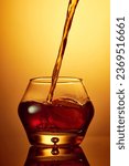 Small photo of Close up photo of splash of pouring to glass whiskey, rum, brandy or gin with ice cube over golden gradient background. Concept of alcohol, strong spirits, drinks, liquid, nightlife, restaurant. Ad