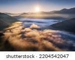 Mountains in low clouds at sunrise in autumn. Aerial view of mountain peak in fog in fall. Beautiful landscape with rocks, forest, sun, purple sky. Top view of mountain valley in clouds. Foggy hills